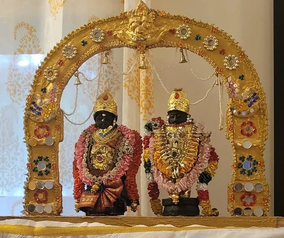 Two dolls of a deity couple in gold ornaments and flower garlands standing beneath a golden arch set with mirrors and beads