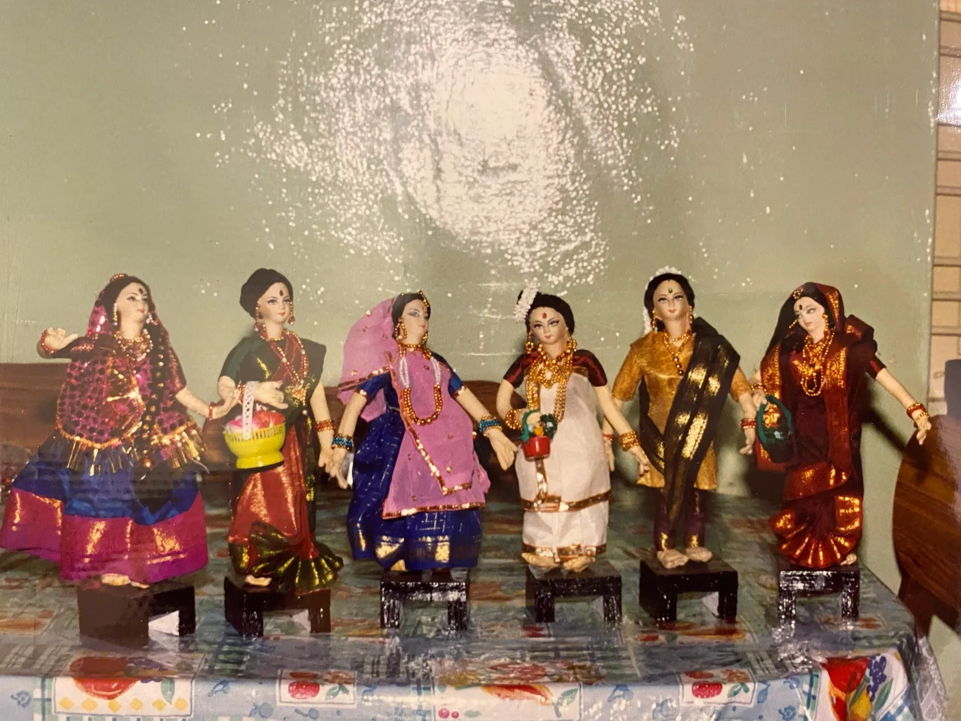 Seven cloth dolls of women in bright silk saris seated in a row on small wooden stools, photographed in 1997