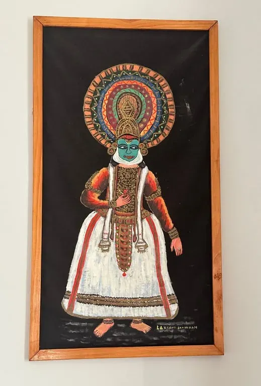 Fabric painting of a Kathakali dancer with a green face, red and white costume and a large headdress, framed on black