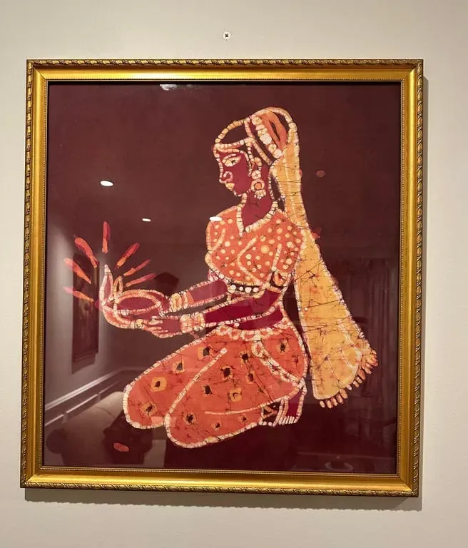 Framed fabric painting of a kneeling woman in an orange sari holding a lamp, on maroon