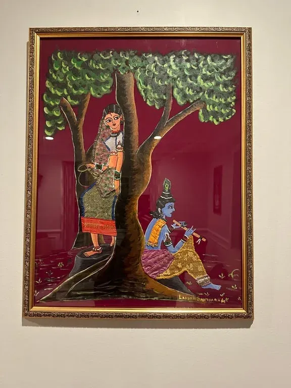 Framed painting of Krishna playing the flute beneath a green tree while a woman leans against its trunk, on a maroon ground
