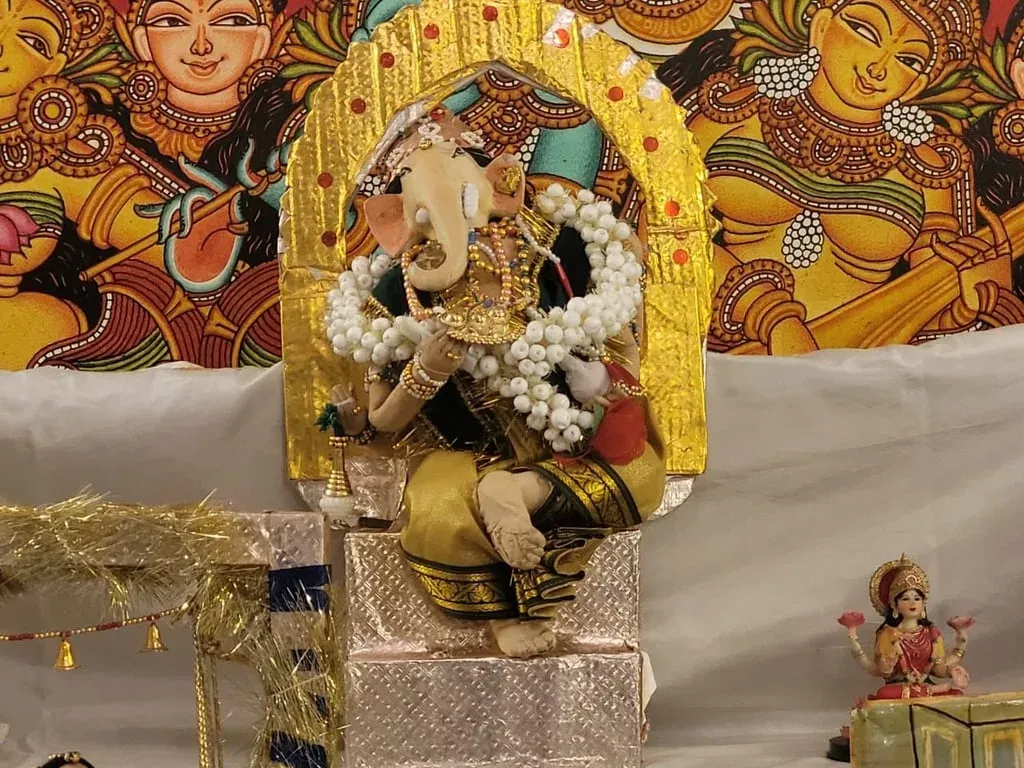 A cloth doll of Ganesha seated beneath a golden arch, with a Kerala mural style painted backdrop