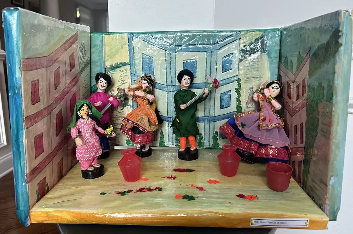 Painted box scene for Holi: dolls throwing colours in a courtyard with blue-washed houses behind