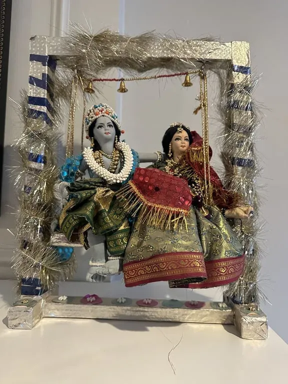 Radha and Krishna seated together on a swing framed in silver tinsel, in green and red brocade