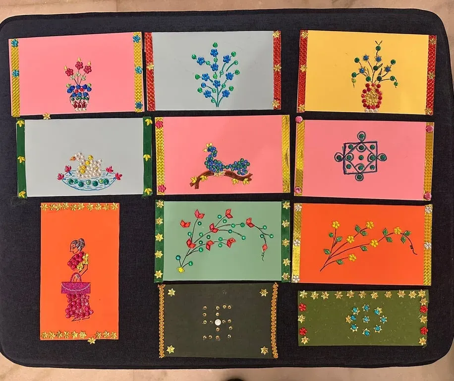 Twelve greeting cards decorated with coloured crystals in the shapes of flowers, plants and kolams, laid out on a black board