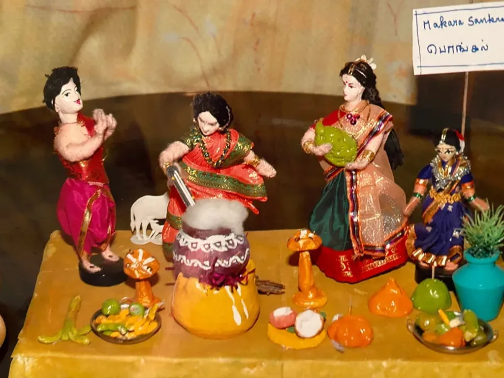 Doll scene of women boiling pongal in a pot, with a cow, lamps and a Tamil label reading Makara Sankaranthi