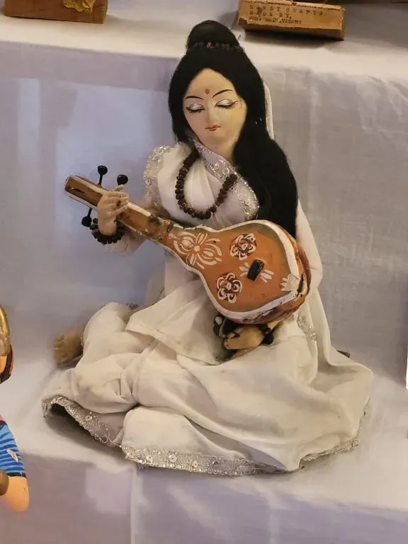 Meera seated in white playing a tanpura: a cloth doll with long black hair and a painted instrument