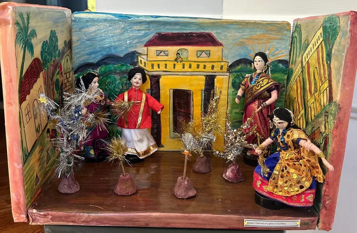 Painted box scene for Diwali: a yellow house hung with lamps and dolls holding sparklers in the courtyard