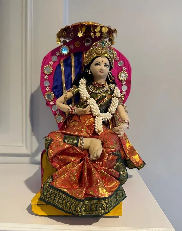 A seated goddess doll in red and gold silk with a crown, a garland and a pink halo behind her