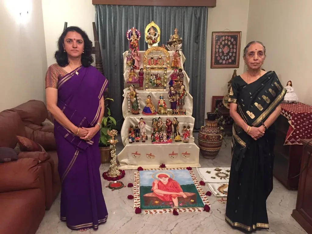 Two women in silk saris standing either side of a tiered golu display with a rangoli on the floor in front, 2017