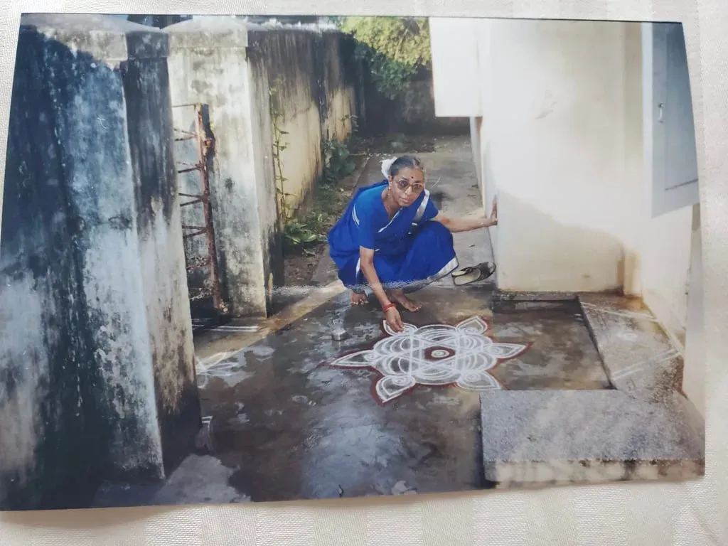 A woman in a blue sari crouching to draw a kolam at a threshold, photographed from the doorway