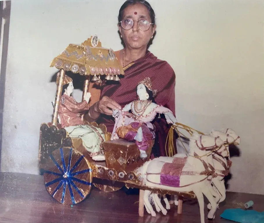 A young Lakshmi Santhanam in a maroon sari beside her doll of the Geetha Upadesam scene: a canopied chariot with two figures, drawn by two white horses, an older photograph