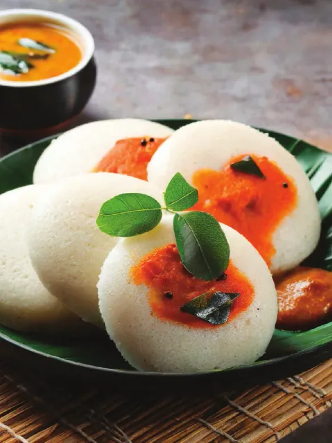 Idlis on a banana leaf with tomato and coconut chutneys, sambar, and a curry-leaf garnish