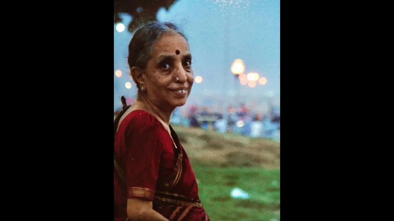 Lakshmi Santhanam in a red silk sari, smiling over her shoulder outdoors at dusk, with lights blurred behind her